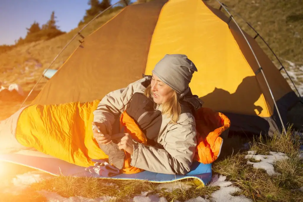 Girl checking her phone by the fire in her sleeping bag - Can I Wash My Down Sleeping Bag