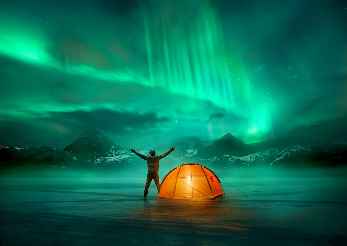 A man camping in wild northern mountains with an illuminated tent viewing a spectacular green northern lights aurora display.