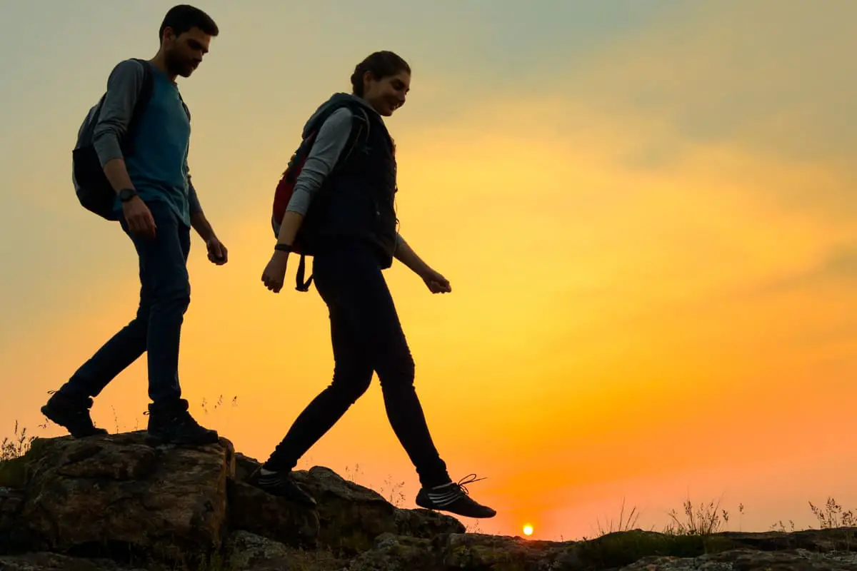 Couple of Young Happy Travelers Hiking with Backpacks on the Beautiful Rocky Trail at Warm Summer Sunset