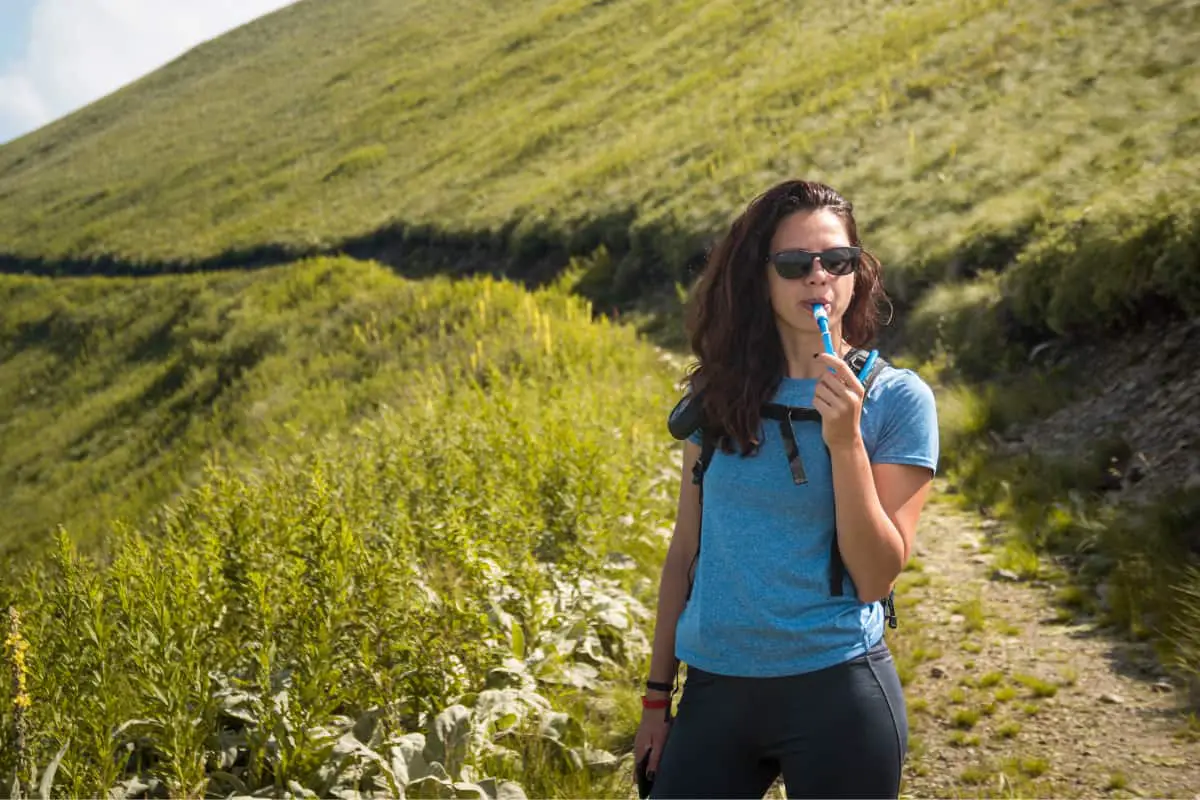 Woman in a blue shirt drinking water from her hydration pack on a trail with grass on the side of a mountain.